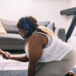 Plus-size woman with headphones kneels on the floor looking at her mobile phone.