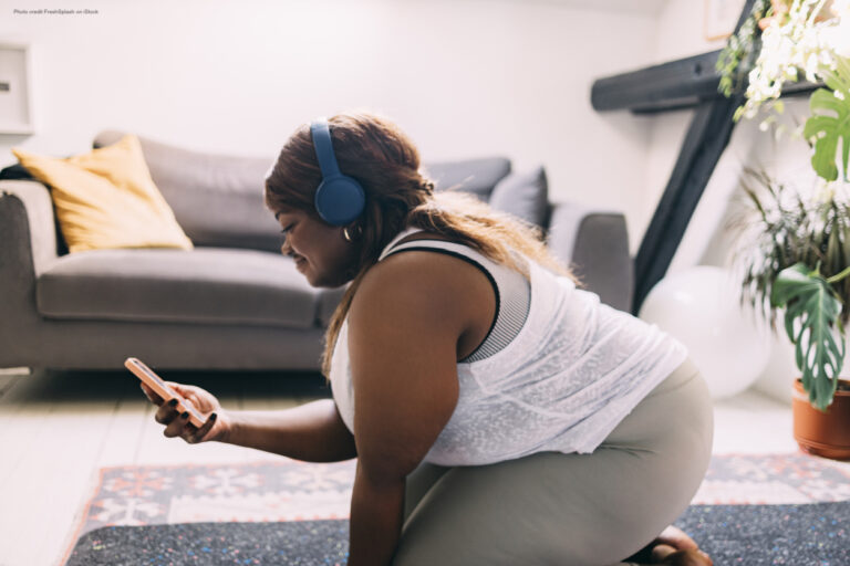 Plus-size woman with headphones kneels on the floor looking at her mobile phone.